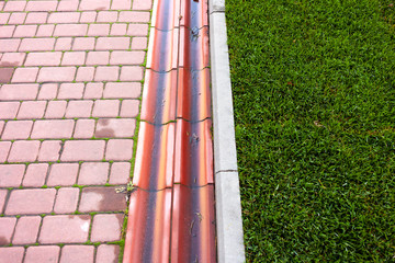 Grid drainage system in the park by the green lawn and footpath of red paving slabs.