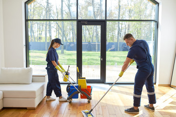 Caucasian janitors in blue uniform washing floor, using detergents, panoramic window background,...