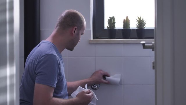 Bald man grabs the last strip of paper from a toilet paper roll. He is getting angry after remaining without toilet paper.