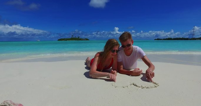 Romantic tourist couple lying on white sand beach drawing a heart with a  clear blue skies in Caribbean - Steady Shot