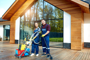 Two caucasian janitors, cleaners stand posing after successful job in big wooden house with panoramic windows. Holding mop, detergents