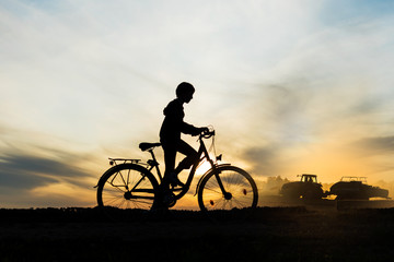 Obraz premium Boy , kid 10 years old riding bike in countryside, tractor working in background, silhouette of riding person and machine at sunset in nature