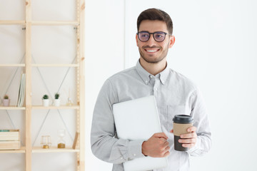Caucasian guy standing in office pressing laptop to his chest and holding coffee cup, dreaming...