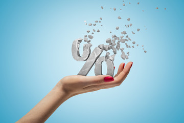 Closeup of woman's hand holding percent sign which is breaking into small pieces that are flying away on light blue background.