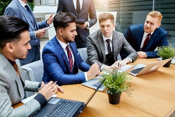 Young men gathered to discuss new project, use laptop, sit on table, young businessmen in office wearing tuxedo