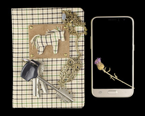 A handmade notebook on which lies a bunch of keys and a pendant in the form of an owl, and next to the smartphone on which lies the thistle flower