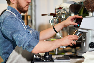 Gentle hands of beard young barista hold metal pot, worm steamed milk, pour ground coffee, push button of coffee machine in coffee bar, professional concept