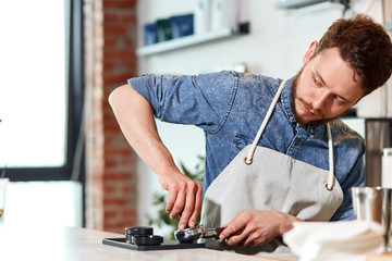 Professional young man with thick stylish beard, wears blue jeans shirt and neat apron, presses coffee using tamper, looks aside at customers, working hard in coffee house
