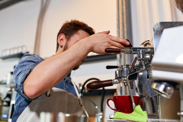 Young bearded man stands near professional grinder, keeps handle, watches coffee pouring process, prepares fresh aromatic cappuccino for customer in modern restaurant