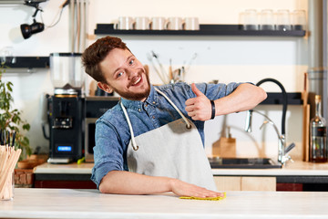 cheerful. optimistic man with thick beard, wears jeans shirt and neat apron, showa thumb up gesture, gradfully smiles, looks at camera with happy face, in brightly lighted room