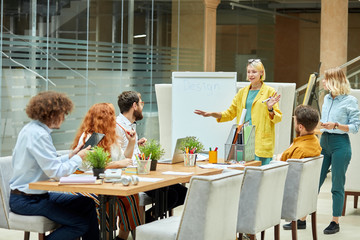 Joyful delighted lady dressed in casual outfit, spreads hands sideways, tells theme of meeting, curious coworkers listen to speaker attentively. Designers meeting concept