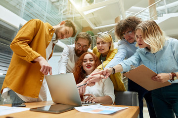 Bottom shot of cheerful people with charming foxy haired female in the middle, stand in modern office, focused in laptop screed, express joyful expression, positive concept