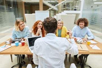 Back of male team leader, sitting at table in front of young talented designers, gestures passionately, talks to pleasant colleagues at daily meeting in comfortable office