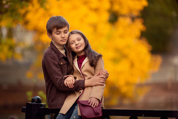 Little Loving boy and girl are sitting on a walk in the autumn park with their eyes closed and dreaming of yellow beautiful trees behind them.