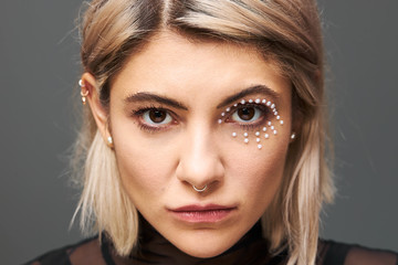 Naklejka premium Close up portrait of beautiful charming young blonde female model with short dyed hair staring right at camera, having mysterious intent gaze, wearing black dress, face piercing and glamorous make up