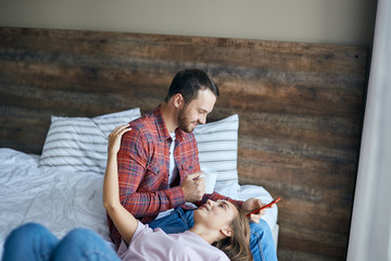 Fototapeta premium romantic couple dressed in casual outfit, resting in bedroom, pretty girl lying on bed, putting head on knees husband, looking up with happy expression, stretches hand ut to hug delighted man