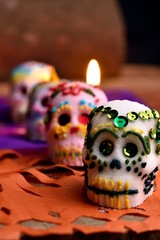 Skulls made by hand, made of sugar, decorated with colored glass and sequins, behind a lit candle. Placed in confetti and wood. Days of the dead Mexican tradition