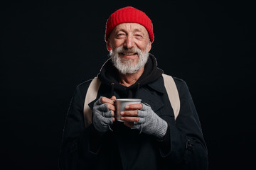 Bearded beggar male smiling, happy even living in street. Cheerful homeless man with mug of hot tea...