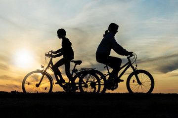 Boy and young girl riding bikes in different directions,  silhouettes of riding persons at sunset in nature