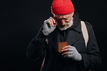 Senior man in black street clothes, red hat and ragged grey gloves look at piece of bread with hope. Crying. Leftover food for all day. Isolated over black background