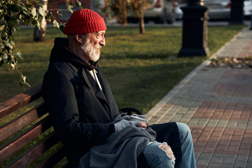 Mature man without shelter sitting in street clothes on bench, without food and money. Drooping man lowered his head down. Side view on homeless person in coat and red cap