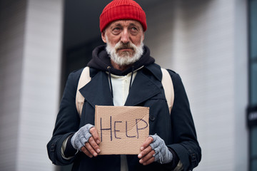 Mature man asking for some help standing on city street. Homeless man in warm coat and red hat