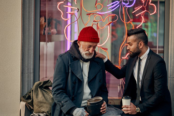 Businessman in tuxedo and homeless person in dirty clothes sitting and talking in the street, look...