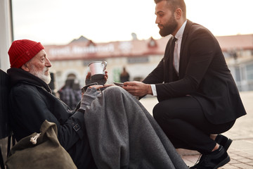 Kind man in suit hunkered down to homeless and give money donation, one dollar bill to beggar male