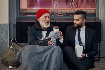 Young beardy man in suit sitting with beggar on floor on street and give cup of coffee. Different...