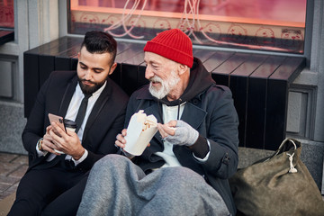 Handsome man in suit show smartphone to beggar man sitting on street