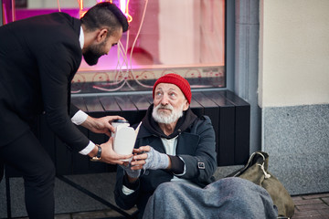 Kind rich man in black tuxedo proposing box filled with warm food and coffee to thankful senior...