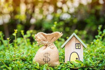 Home model and money bag put on the grass on the sunlight in the public park, Saving money for buy a house or loan for plan business investment of real estate concept.