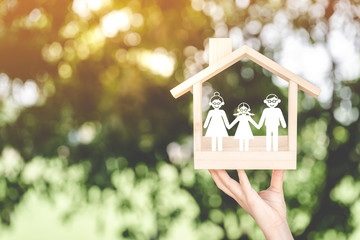 Woman hand holding a wooden home with happy family of paper art is placed inside on nature bokeh in the public park, The saving money for house or real estate owner in the future concept.
