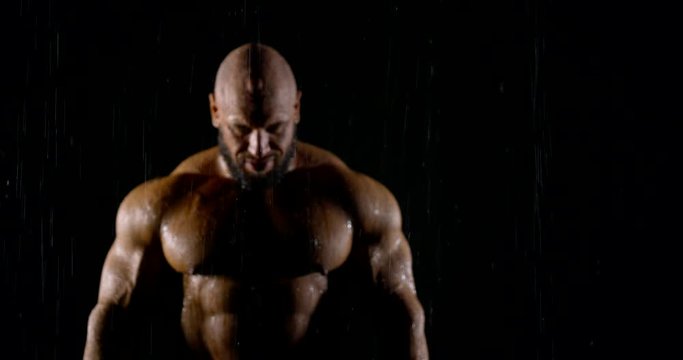 a large muscular athletic bald man with a beard goes to the camera in the rain and gets into focus. drops of water trickle down his body. dark key