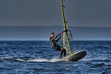 Naklejka premium A male athlete is interested in windsurfing. He moves on a Sailboard on a large lake on an autumn day.