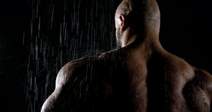 a very large athletic muscular bald man stands in the rain. drops of water trickle down his broad back. dark key
