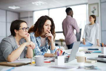 Portrait of two young businesswomen looking at laptop screen and thinking while planning creative...