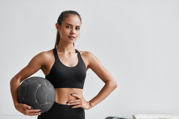 Sportive caucasian cross fit woman with fitness ball stand in gym, look at camera. White wall background