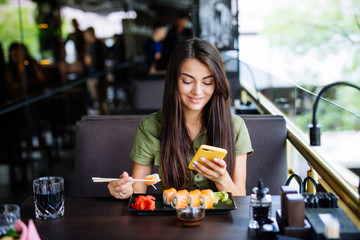 Young woman texting on the phone while eating sushi in a restaurant