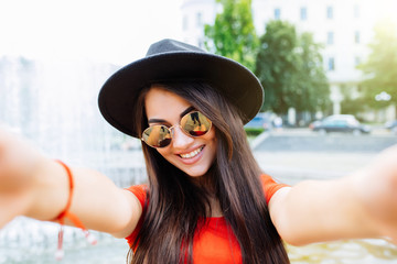 Young woman in hat and sunglasses take selfie on the sunny summer street