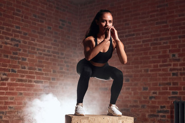 Slim sport woman in sportswear jumping on fit box and do squats. Crossfit, bodybuilding. Brickwall background