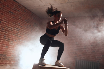 Attractive sporty female with long hair doing squats on wooden box. Process of jumping. Brickwall background