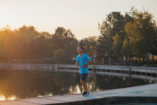 Young man in sportswear jogging by the lake in a city park. Runner training in the park by the water at sunset.
