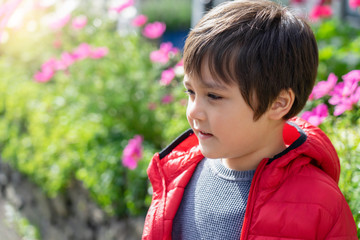 Portrait of Happy young boy standing in the park with blurry flowers background, Active child looking out with smiling face wearing red jacket, Kid having fun playing outdoors in sunny spring