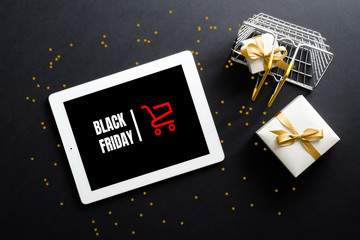 Black Friday sale concept. Tablet with sign "Black Friday" on screen, shopping basket, gift box, confetti over black background. Flat lay, top view, overhead.