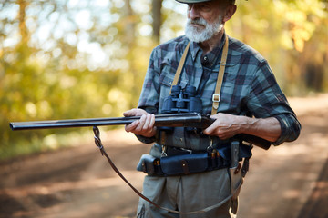 hunter man with grey beard holding gun to hunt on birds, ready to shoot. Forest background