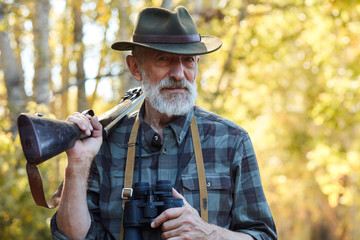 Satisfied man with grey beard look at camera, stand holding binoculars in one hand, shotgun on shoulders. Forest,autumn trees background, sunny day