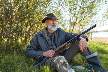Bearded male with shotgun sitting on grass, have rest after hunting, wearing hat, coat and rubber boots