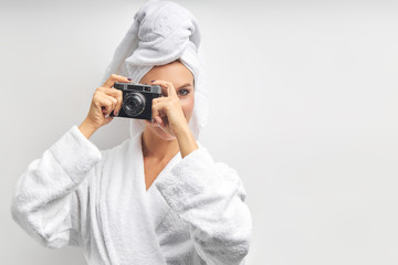 Caucasian pretty woman stand taking photo with photo camera, dressed in white bathrobe and towel on head, look at camera with one eye. White isolated background