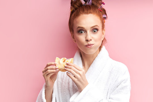 How delicious it is. Portrait of beautiful girl with sweet tasty donut, eating after shower, dressed in white bathrobe, curlers on hair. Isolated pink background